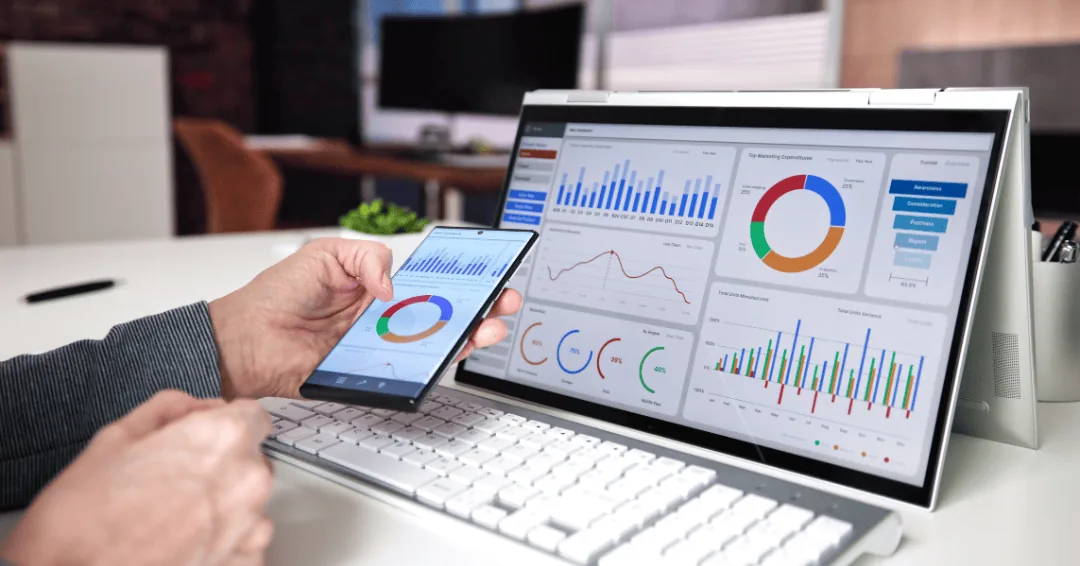 Person holding a phone with charts beside a laptop showing a marketing analytics dashboard