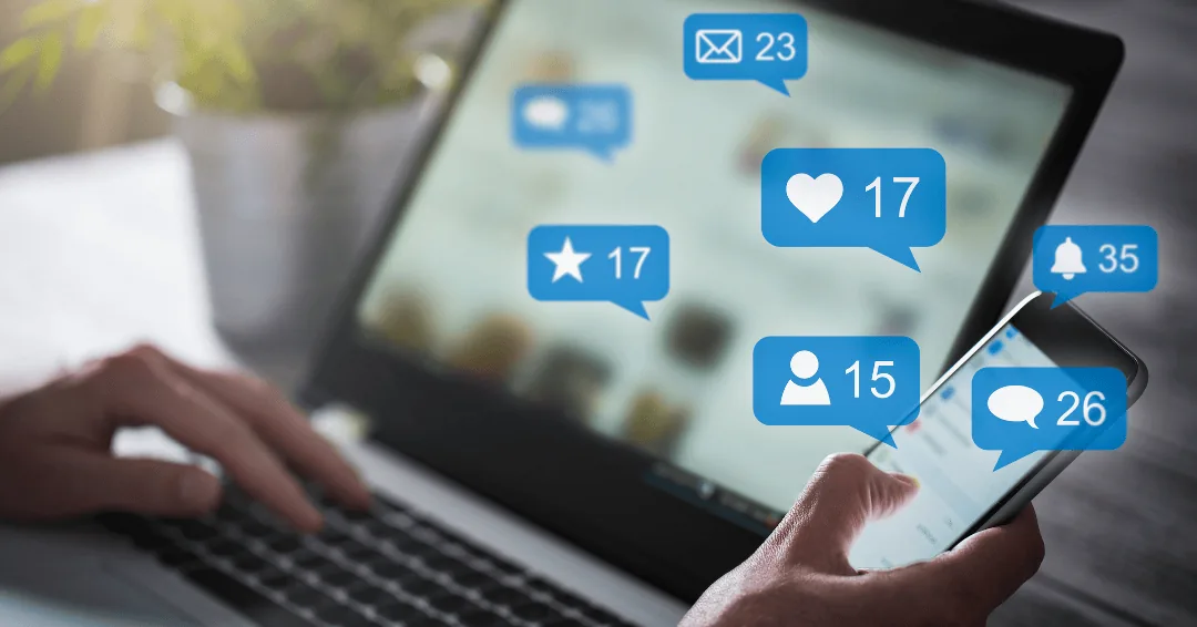 Hands using a laptop and smartphone with social media notification icons for likes, messages and followers